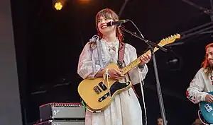 Teasdale in a white dress standing onstage with a guitar, smiling and looking to the left