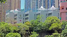 A white concrete building with "Glenealy School" and some Chinese writing and an ESF logo on the front, seen from below and at a distance. There are green treetops below it and portions of taller buildings in the background.