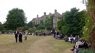 extensive gardens with English country house in the background; people in evening dress stroll on the lawns