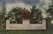 Picture postcard of a large bronze statue of two men. The statue is on top of a stone pedestal that is flanked on each side by stone walls with stone benches running along the base of the wall. The pedestal, walls, and benches sit on top of a large stone step. The monument has grass in front of it, and trees behind it. There is printing along the top of the postcard that says "No. 140. Goethe–Schiller Monument. Washington Park. Milwaukee, Wis."