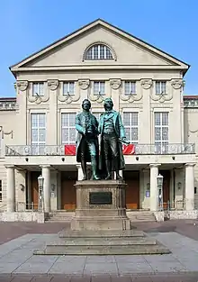 Photo of a statue of two men in front of a large stone building with a colonnade at the entrance, many rectangular windows and carved decorations at roof level