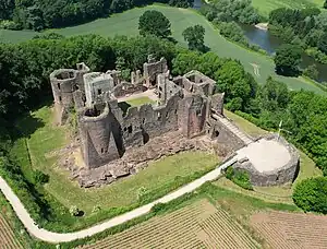 An aerial view of a ruined castle surrounded by trees. Inside the walls is a delapidated keep made from a lighter stone.