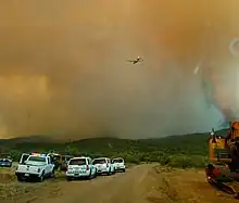 First responders vehicles along a road in the Bradshaw Mountains. Smoke from the Goodwin Fire billows over the road and a firefighting aircraft.