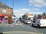 The junction of High Street and Church Road. A terrace of stone fronted buildings, with a curved corner building fronted in white, from the mid-late 19th century. The modern shop fronts are not in keeping the original style of the buildings. It is set back from the road by modern paving. The far section of the building is the old fire station building (formerly Urban District offices) are .