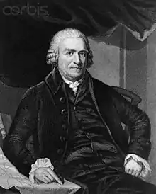 An older man, seated, with a hint of a smile. He has white hair and is wearing a dark suit. He is pointing to a document on a table.