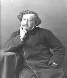 A man with medium-length hair rests against a table, a finger pressed against his right cheek.