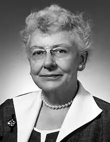 a black-and-white head-and-shoulders portrait of a smiling grey-haired woman wearing a smart dress, a pearl necklace and earring, a small broach and rimless pince-nez spectacles.