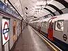 The interior of a building with a rounded, white roof, white walls, grey flooring, and a train on the right carrying passengers