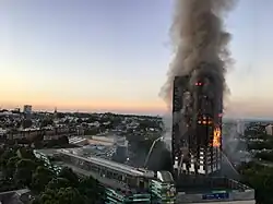 A tower block (Grenfell Tower) burning on nearly all floors with large amounts of smoke rising, and water being sprayed at the building from firefighters.