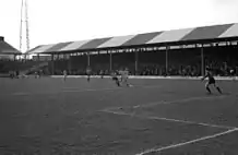 A black-and-white photograph of a football match in progress in front of a modest, single-tiered, British-style grandstand