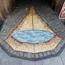 On Peter Street in Dundee, there is a mosaic on the ground commemorating Grisell Jaffray, the last woman to be burnt as a witch there. There are two elements to the mosaic, one representing fire and the other representing water. This image is of blue and gold mosaic tiles surrounded by cobble stones.