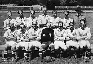 An association football team poses for a formative black-and-white photograph. A row of seven men sits on a bench, all wearing light-coloured shirts and white shorts apart from the player in the centre, who wears black. A football rests on the ground between his feet. Behind the seated row stand seven more men, all but one of whom are wearing the same light-coloured shirts. The exception is a gentleman standing at the end of the row on the viewer's right, who wears a dark double-breasted suit, tie and wide-brimmed fedora hat. All of those present have their arms folded apart from the man in the suit, whose hands are behind his back. In the background a set of goalposts can be seen.