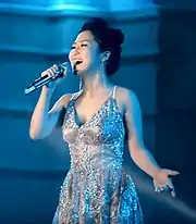 A picture of a brunette woman performing on stage, she is wearing a grey dress decorated with sequins