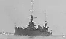 Photograph of a large warship. Along the bottom edge of the photograph, the phrases "HMAS AUSTRALIA" and "BUILT FOR THE AUSTRALIAN NAVY" are written in white. A sailboat and distant land can be seen in the left part of the photo.