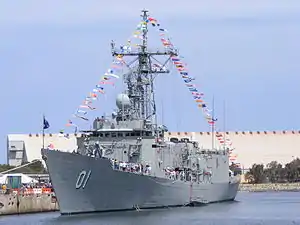 A warship tied up at a wharf. The ship has the number "01" painted near her bow. She is decorated with flags and bunting, and a large number of civilians stand on her decks