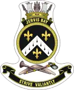 A ship's badge. A naval crown sits on top of a black scroll with "JERVIS BAY" written in gold. This is atop a yellow, rope-patterned ring, which contains a black field. Three gold fleur de lys are positioned at the bottom, upper left, and upper right: the bottom one is separated from the other two by a white chevron containing eight black marks. Below the ring are a stone axe and a nulla nulla sitting on top of a boomerang. At the bottom of the badge is a black scroll with "STRIVE VALIANTLY" written.