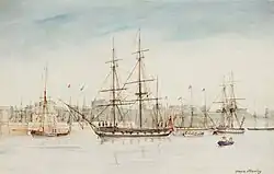 Square-rigged three masted naval sailing ship moored at a port, with other ships and the seawall with buildings behind it in the background. Seen from the water, with a small boat being rowed in the foreground.