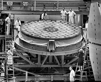 A black and white photograph of a large, flat mirror with a geometric pattern, supported on a metal frame with several people surrounding it.