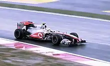 A figure, wearing a yellow and white helmet design, is driving a Formula One car that is of a silver and red colour scheme.