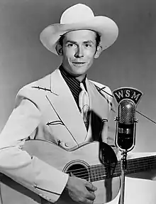 A photograph of a young man wearing a cowboy hat, holding a guitar and standing next to a microphone