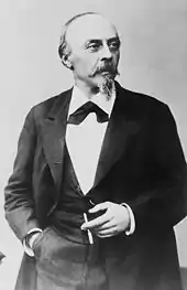  A balding man with a pointed beard, in formal dress clothes and holding a cigar in his left hand