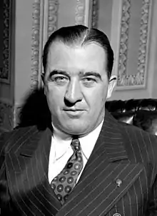 A man in his fifties, with thinning black hair, sitting at a desk, face-forward, wearing a suit