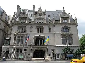 View of the main entrance as seen from across 79th Street. There is a window and fence to the left of the main entrance and shrubbery to the right. Above the main entrance are two stories of windows, housed in either square recesses or semi-elliptical and fully Gothic arches. The house contains a double-height slate mansard roof with dormer windows projecting from it.