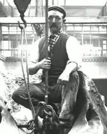 Man wearing working clothes being winched up in a galleried hall, while sitting on top of a sperm whale skeleton