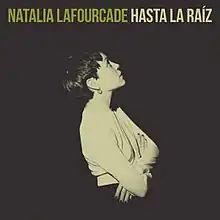 A sepia-toned picture depicting a woman wearing a white shirt looking up to her right. Above her, the words "Natalia Lafourcade" and "Hasta la Raíz" are placed in all caps.