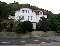 A road and behind it, at a somewhat higher level, a large, three-storey white house with a red roof; a wooded hill is in the background. The main house has an extension to the left, and a separate three-storey wing leading off to the right; the colour of the road surface changes abruptly at the point where the main house ends and the wing to the right begins.