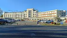 a large brick and concrete building stretches out across the photo about 6 stories high, cars and car parks and bare tress in the front. the sky is blue.