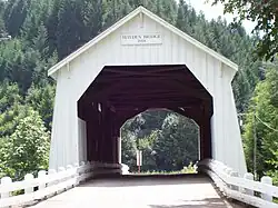 A narrow road leads through a white covered bridge toward a forested hill. The bridge is about 25 feet wide, 25 feet tall to the peak of its roof, and 100 feet long. White guardrails line both sides of the road and the floor of the bridge. A sign above the bridge portal reads, "Hayden Bridge 1918".