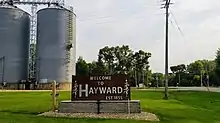 sign reading "welcome to Hayward, established 1855". In the background, grain silos and trees are visible.