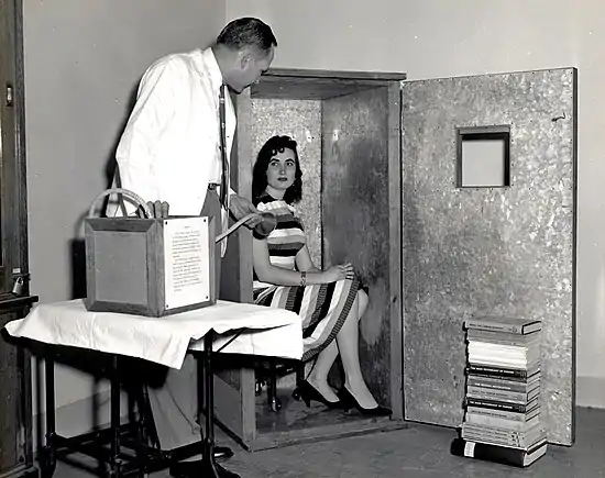 photograph of a woman, sitting on a chair which is in a large, wardrobe-sized box, the open door of which has a small window. A man stands nearby, holding a breathing apparatus.