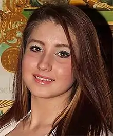 A young Caucasian woman with elbow length straight auburn hair, wearing some lipstick and mascara, looks at the camera, smiling, her head cocked slightly to the right. Behind her is a wall with some patterned wallpaper; the shadow of her head is on it displaced to the right, as well.