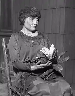 A woman with full dark hair and wearing a long dark dress, her face in partial profile, sits in a simple wooden chair. A locket hangs from a slender chain around her neck; in her hands is a magnolia, its large white flower surrounded by dark leaves.