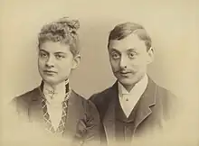 A charming and iconic period photo of a young couple around 1900. The lady has her hair swept upwards and is wearing a high collar fastened with a jewel. The gentleman sports a gallant moustache neatly trimmed and is also wearing a high collar.