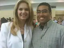 A woman with light skin and blonde hair stands next to a man with dark hair, dark skin, and glasses