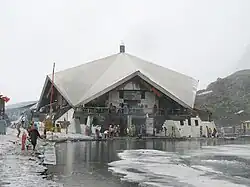 A stone building is surrounded by partially frozen ponds. Pilgrims can be seen on the paths