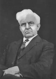 Posed photograph of a white-haired old man, seated, wearing a dark suit