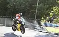 Hervé Gantner wearing a orange high-visibility vest riding a motorcycle on Ballaugh Bridge