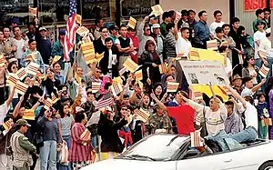 Demonstrators on the sidewalk waving South Vietnamese and U.S. flags. Some are passing by in a car in the foreground.