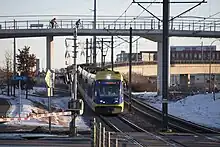 Yellow and blue light rail train travels downhill across a grade crossing; a pedestrian bridge is behind