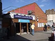 A red-bricked building with a rectangular, blue sign reading "HIGHBURY & ISLINGTON STATION" in white letters all under a blue sky with white clouds