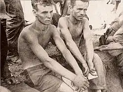 Two men without shirts on sit surrounded by soldiers