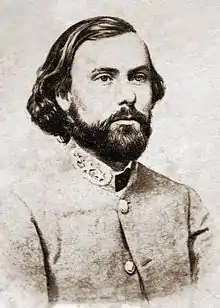 Photo shows a stern, bearded man with long hair wearing a gray military uniform with general's stars on the collar.