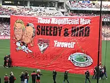 A red banner featuring drawings of former Essendon player James Hird and former coach Kevin Sheedy