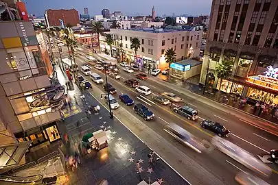 Downward view on a well-trafficked street surrounded by mid-rise buildings. On one side of the street are ceremonial stars embedded in a polished sidewalk.