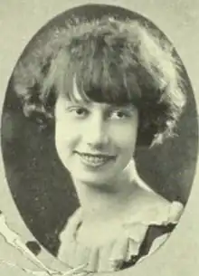 Smiling young white woman with cropped dark hair, wearing a dress or blouse with a scooped and ruffled neckline