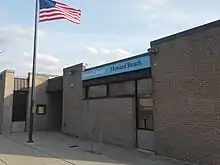 A one-story brick building in a 1960s architectural style stands with a waving American flag before it.